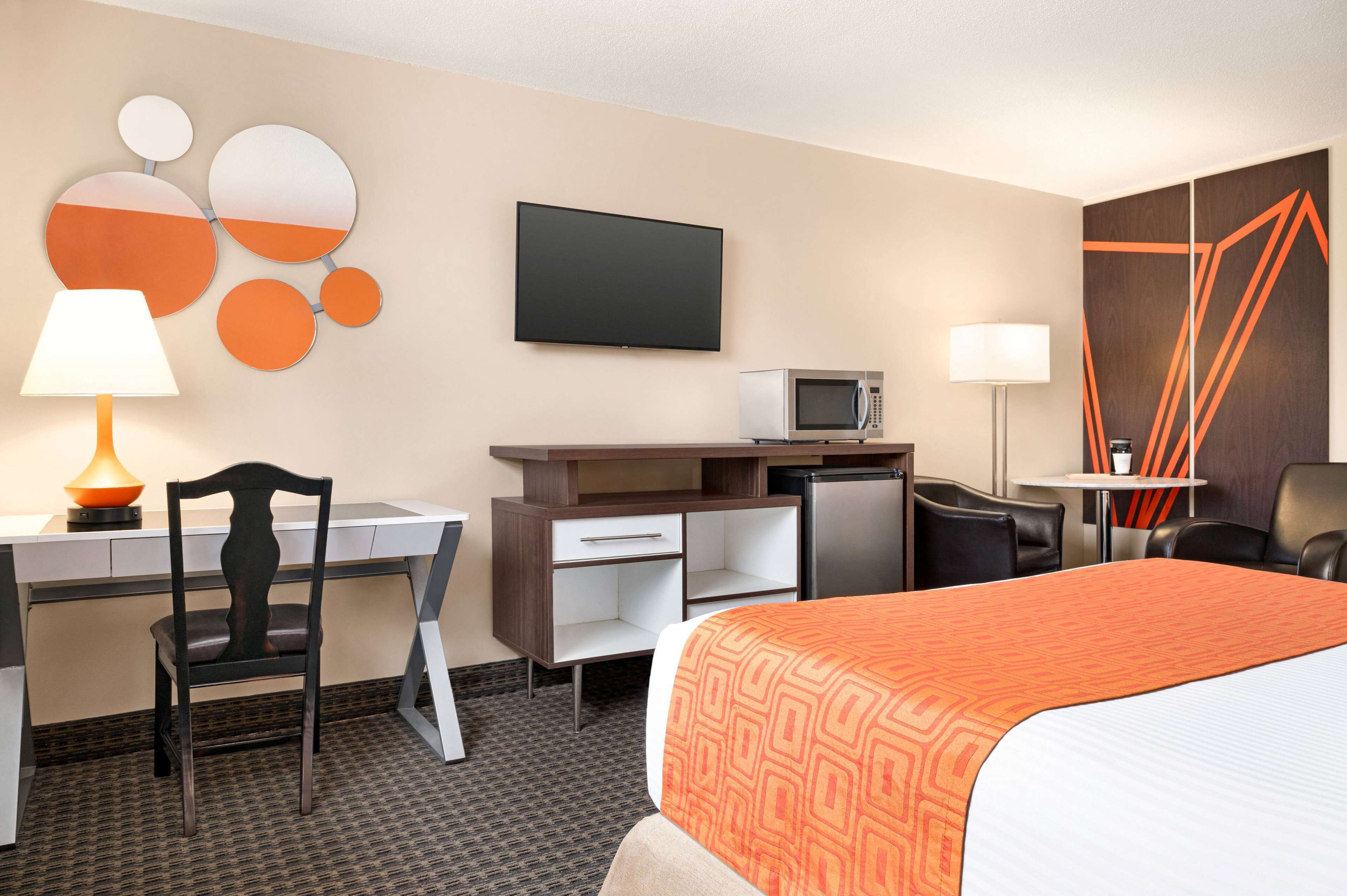 A hotel room with a bed, desk, chair, TV, microwave, and a small table with a lamp.