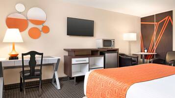 A hotel room with a bed, desk, chair, TV, microwave, and a small table with a lamp.