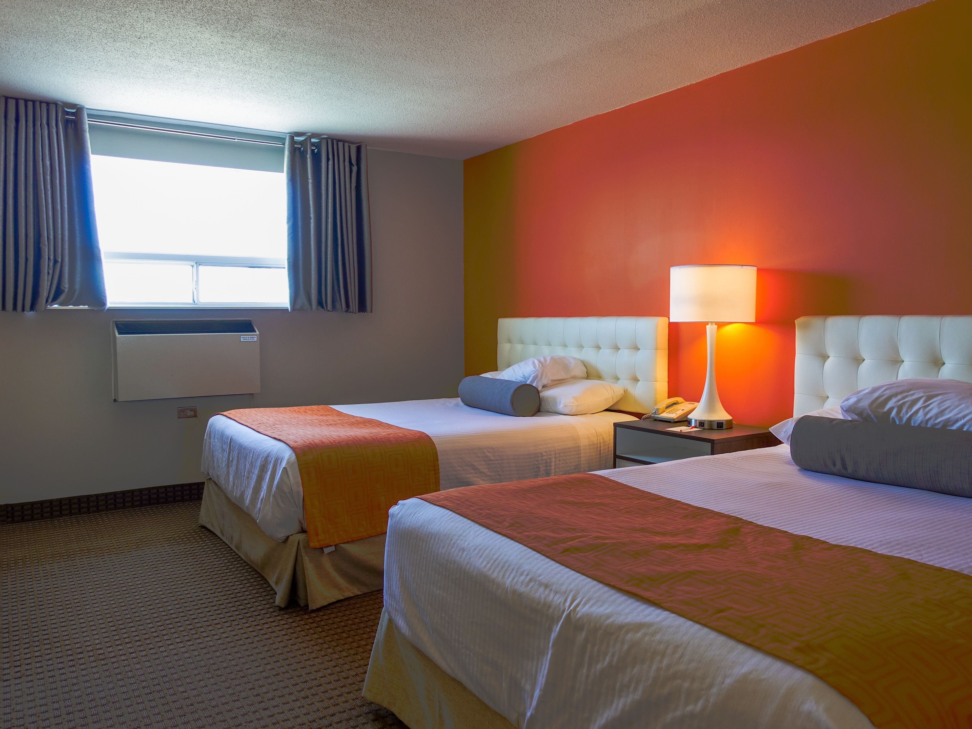 A hotel room with two beds, a window with curtains, a bedside lamp, and a wall heater.