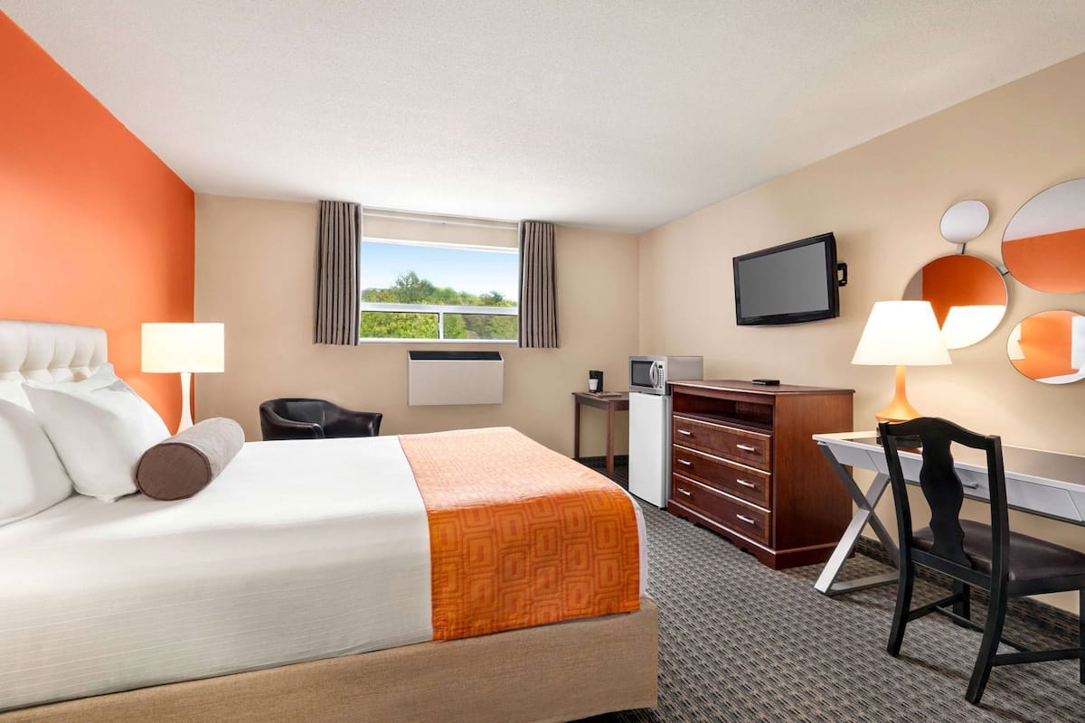 Una habitación de hotel con cama, escritorio, silla, televisor y ventana con cortinas.