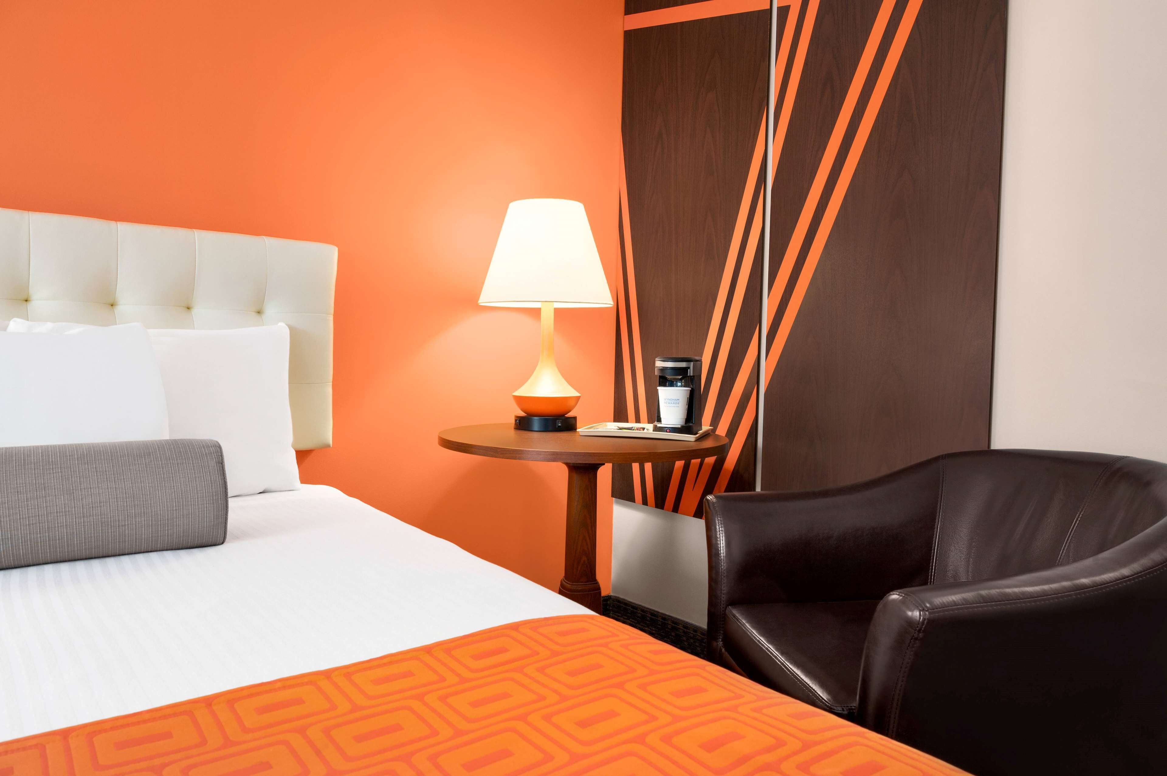 Habitación de hotel con cama blanca, pared de acento naranja, mesa redonda de madera con lámpara, sillón de cuero negro y panel de madera con líneas anaranjadas.