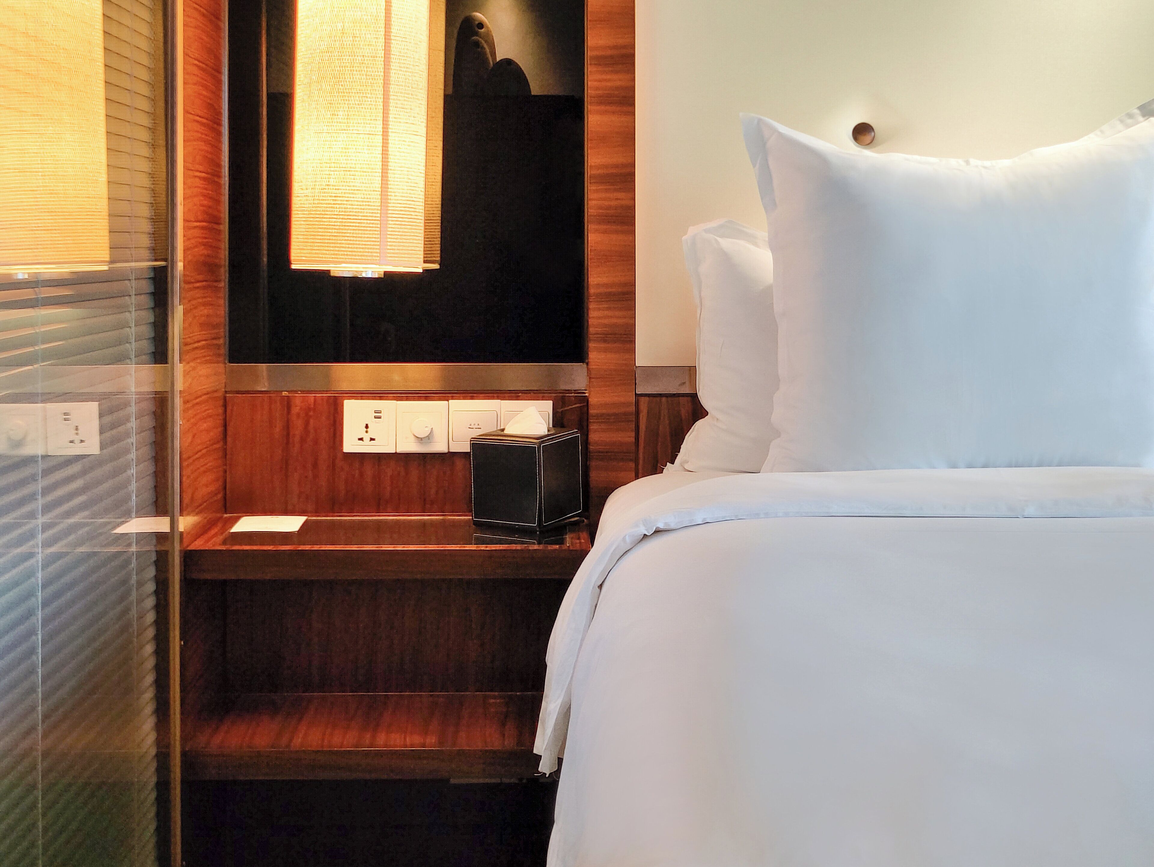 A hotel room with a bed, a bedside table with a lamp, a mirror, and a window with blinds.