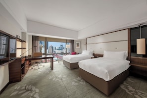 Ein Hotelzimmer mit zwei Betten, einem Schreibtisch, einem Fernseher und einem großen Fenster mit Blick auf die Stadt.