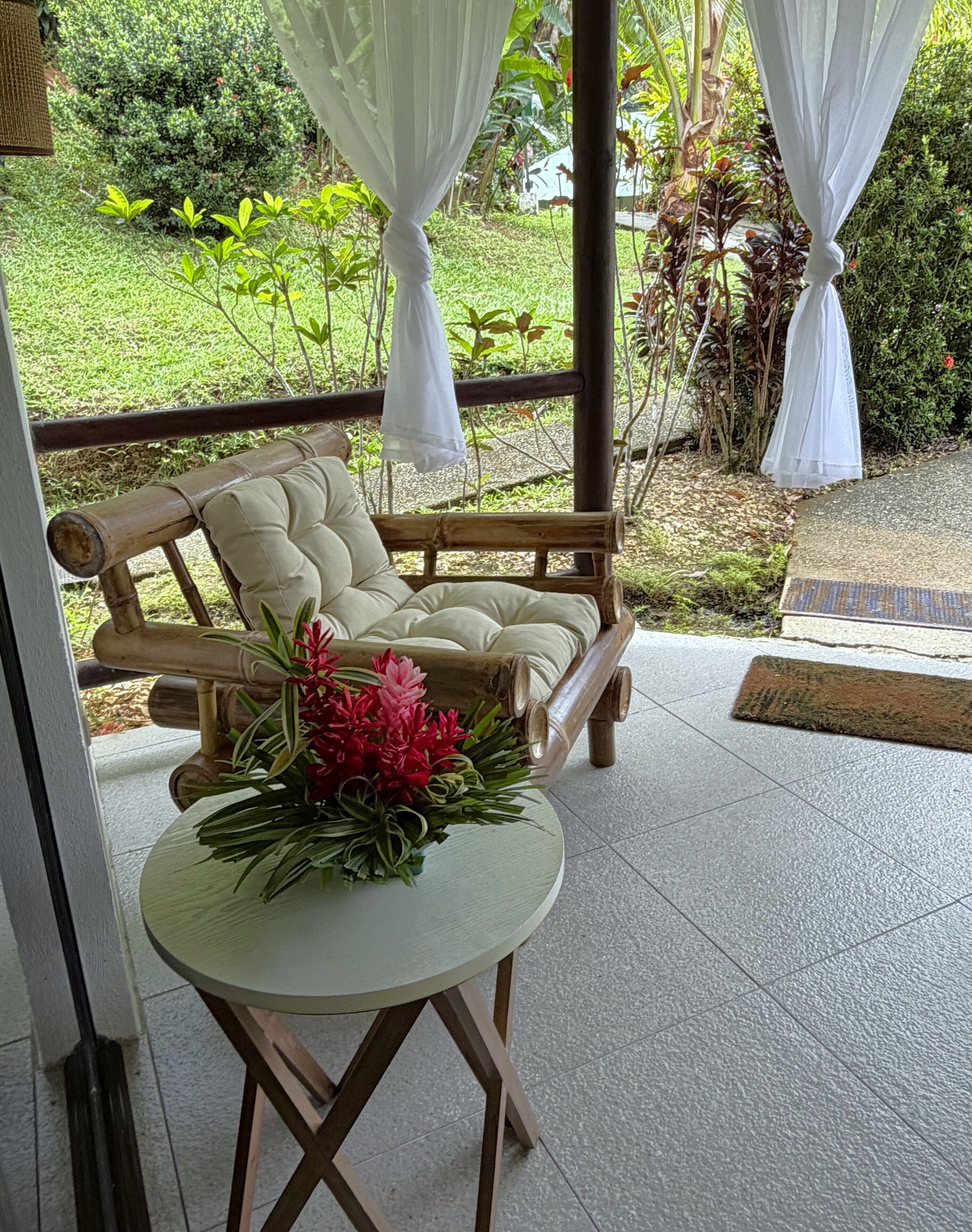 A wooden bench with a cushion, a small round table with a bouquet of flowers, and a view of a garden with greenery and a pathway.