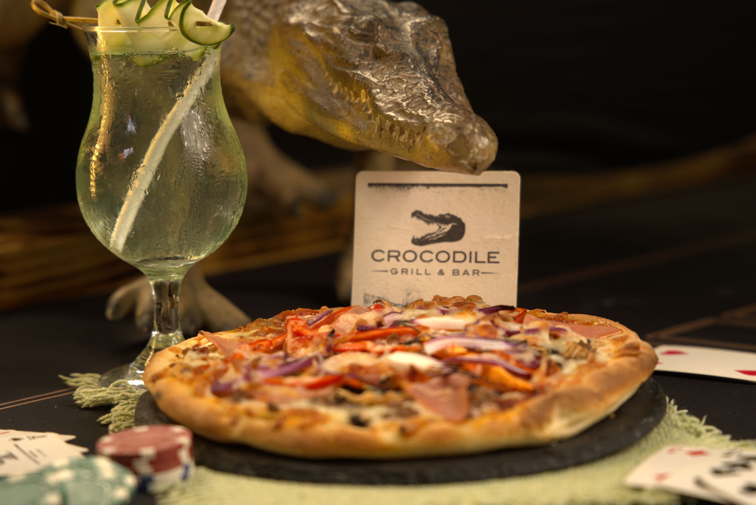 A pizza with various toppings, a cocktail glass, and a crocodile head decoration.