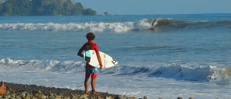 A person standing on a pebbled beach holding a surfboard, with waves in the background and a distant island.