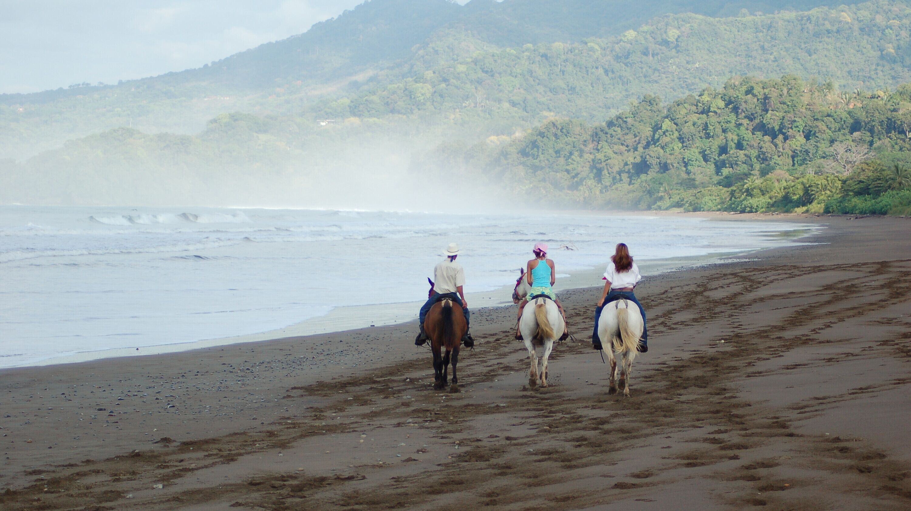 Tres personas montando a caballo por una playa, con una ladera boscosa al fondo.