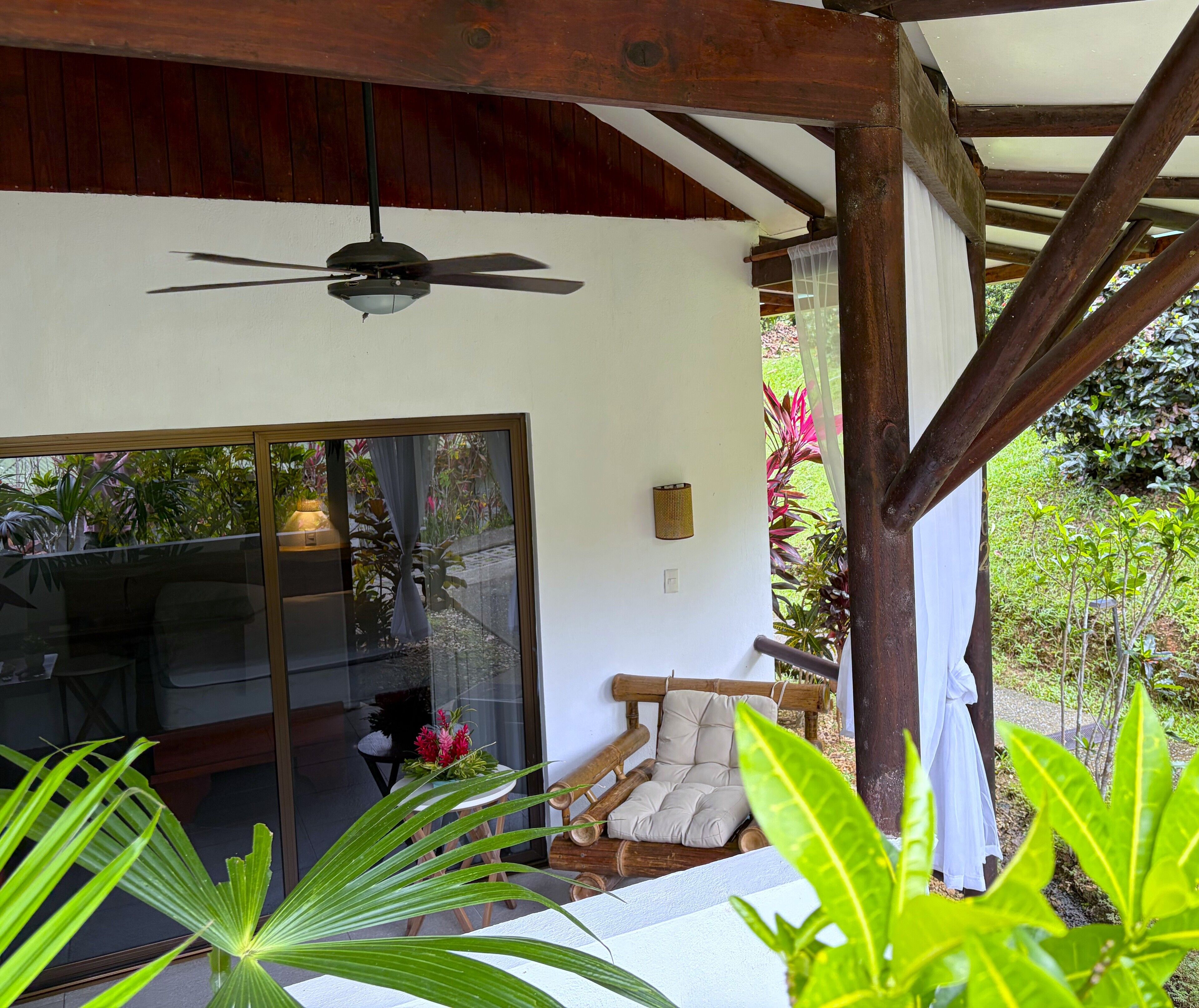 Une pièce dotée d’un ventilateur de plafond, d’une porte-fenêtre coulissante donnant sur un jardin, d’un banc en bois et d’une table avec des chaises.