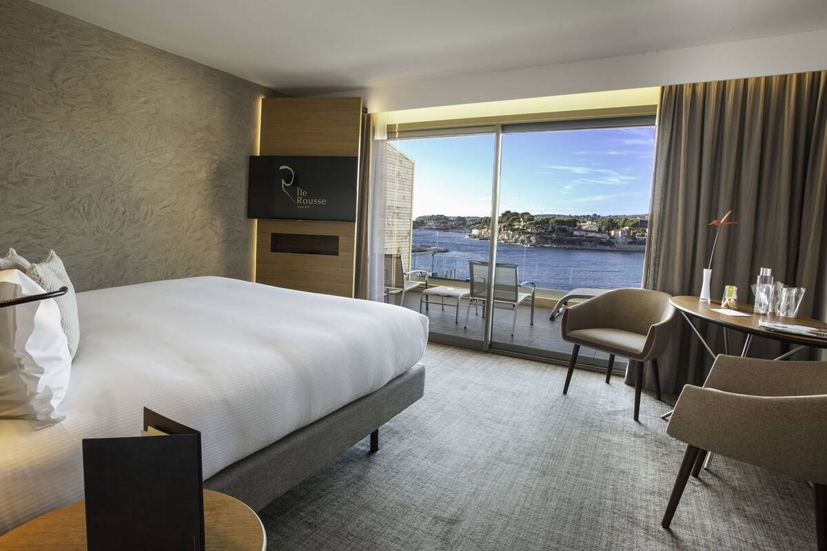 A hotel room with a bed, a desk, a chair, a TV, and a view of the sea.