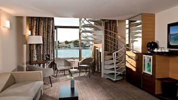 A hotel room with a spiral staircase, a sofa, a small table, a chair, a desk, and a view of the sea.