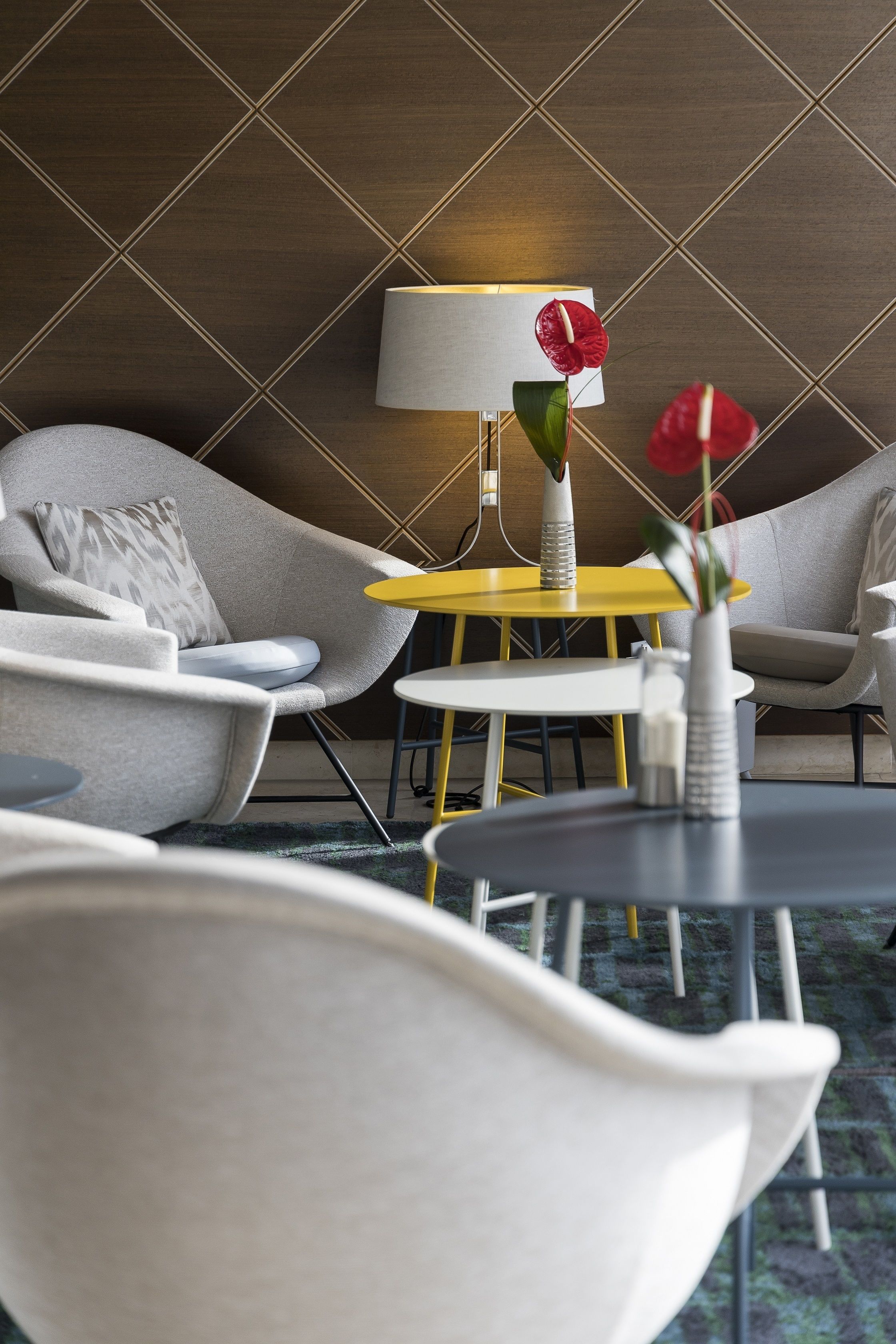 Una habitación de hotel moderna con una pared de diseño geométrico, un sofá gris con un cojín estampado, una mesita auxiliar amarilla, una mesa redonda blanca con un jarrón y flores rojas, y una lámpara con pantalla blanca.