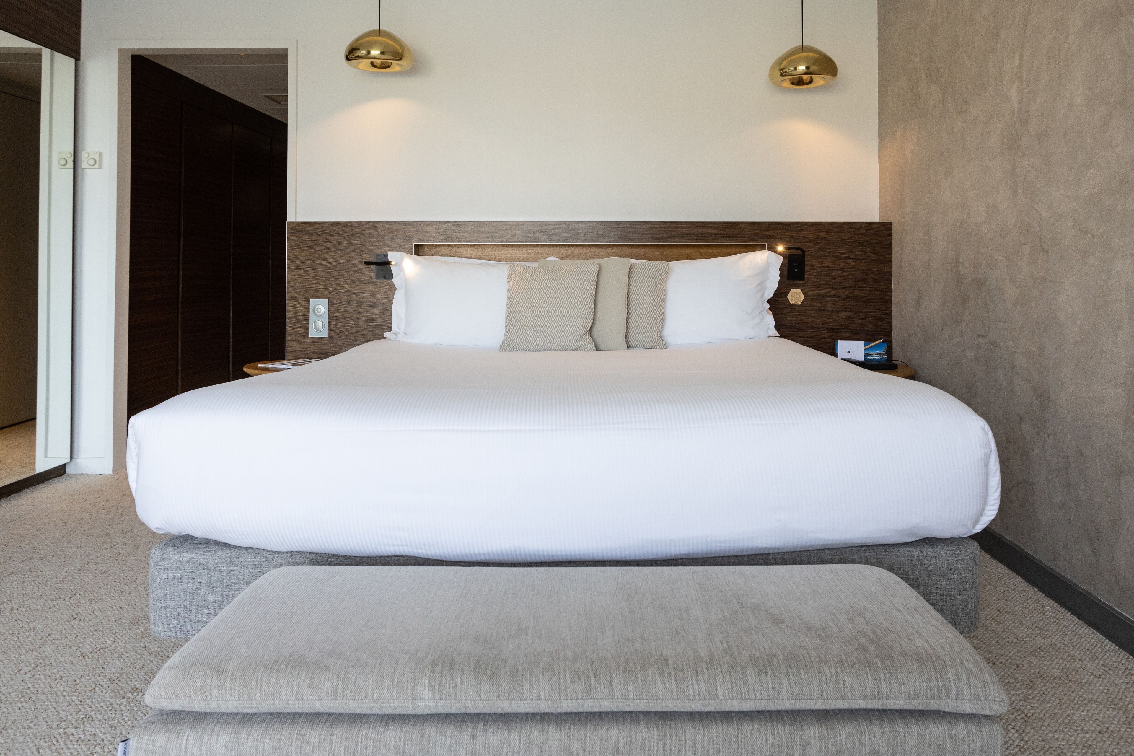 Een groot bed met een houten hoofdbord, twee bedlampen en een grijze poef.