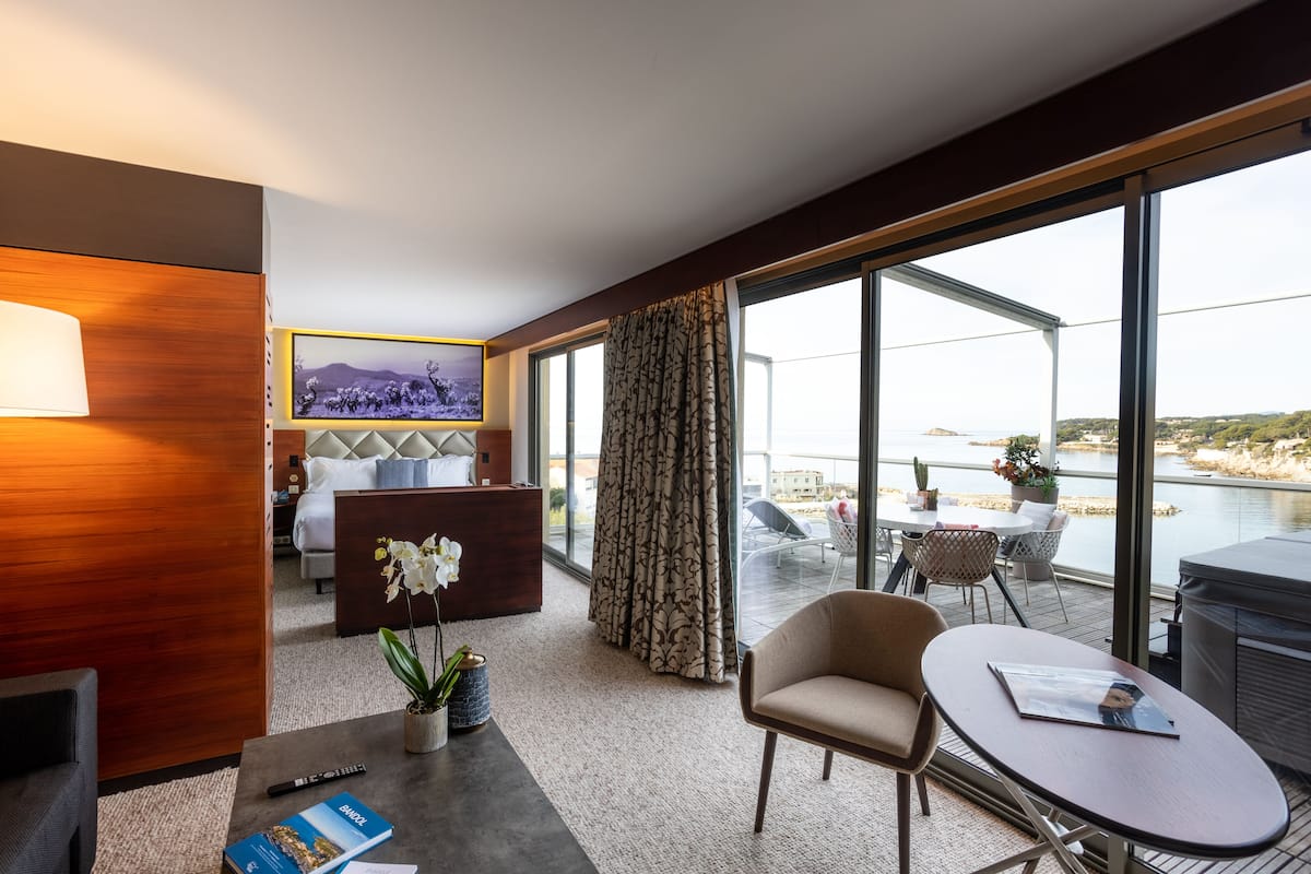 A hotel room with a large window, a bed, a desk, and a seating area with a view of the water.