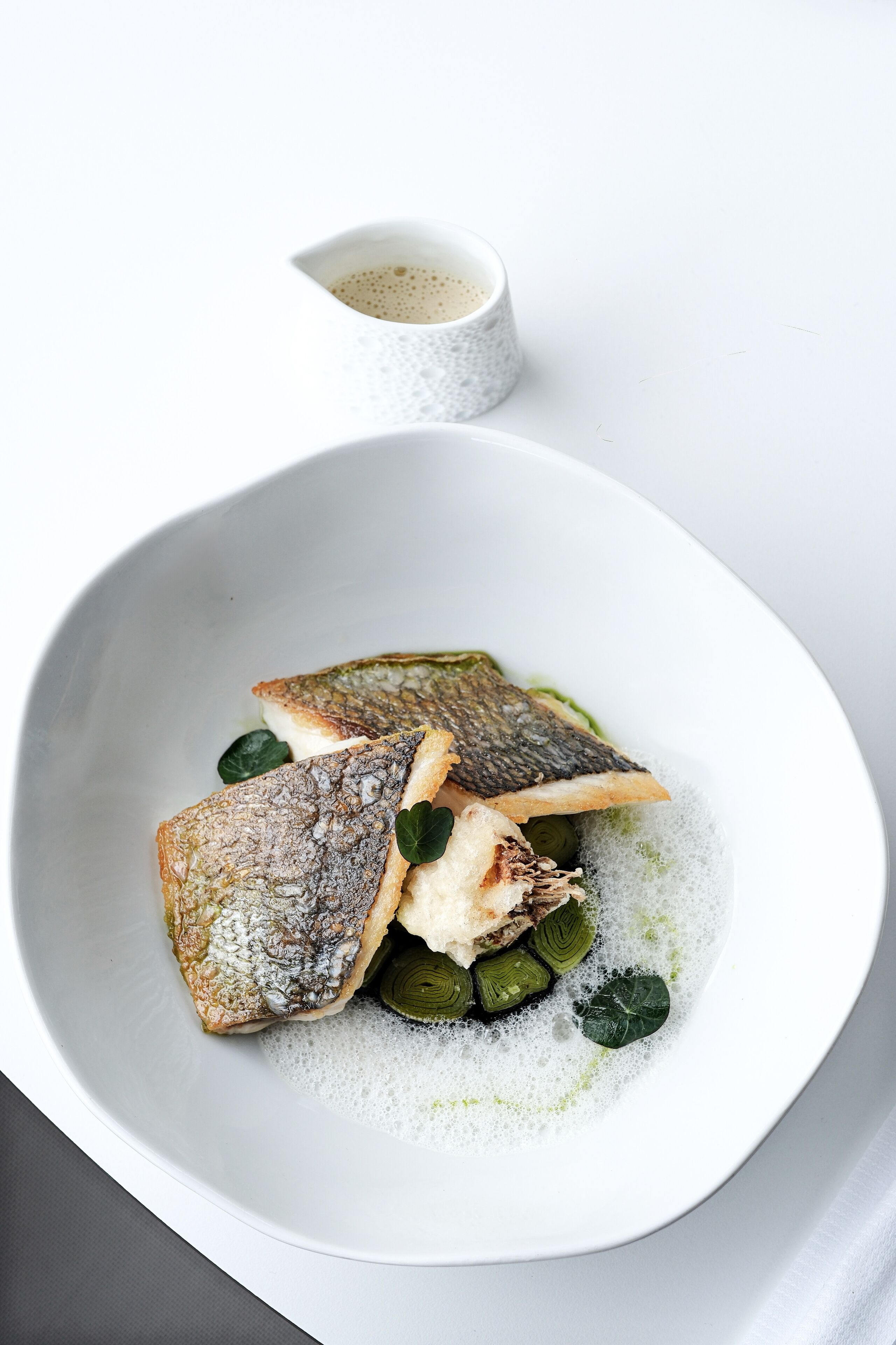 A white plate with fish, greens, and a small cup of sauce.