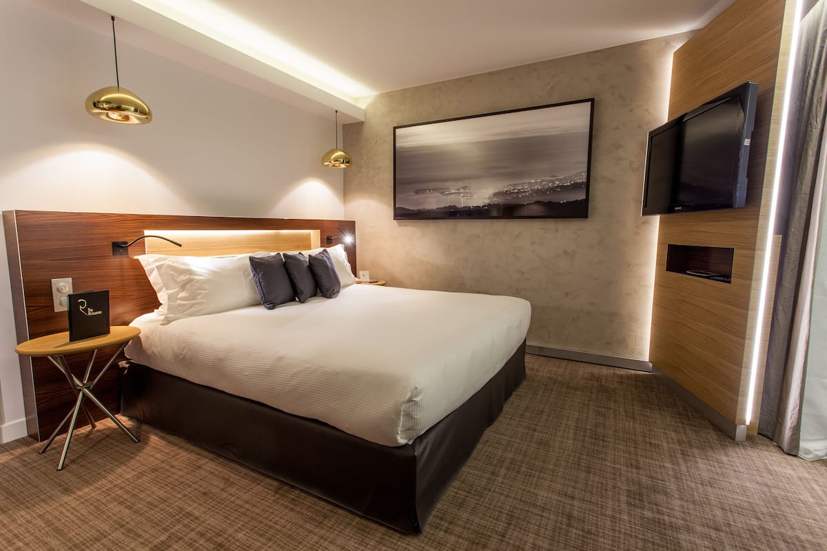 A modern hotel room with a large bed, a flat-screen TV, a painting on the wall, and a small table with a lamp.
