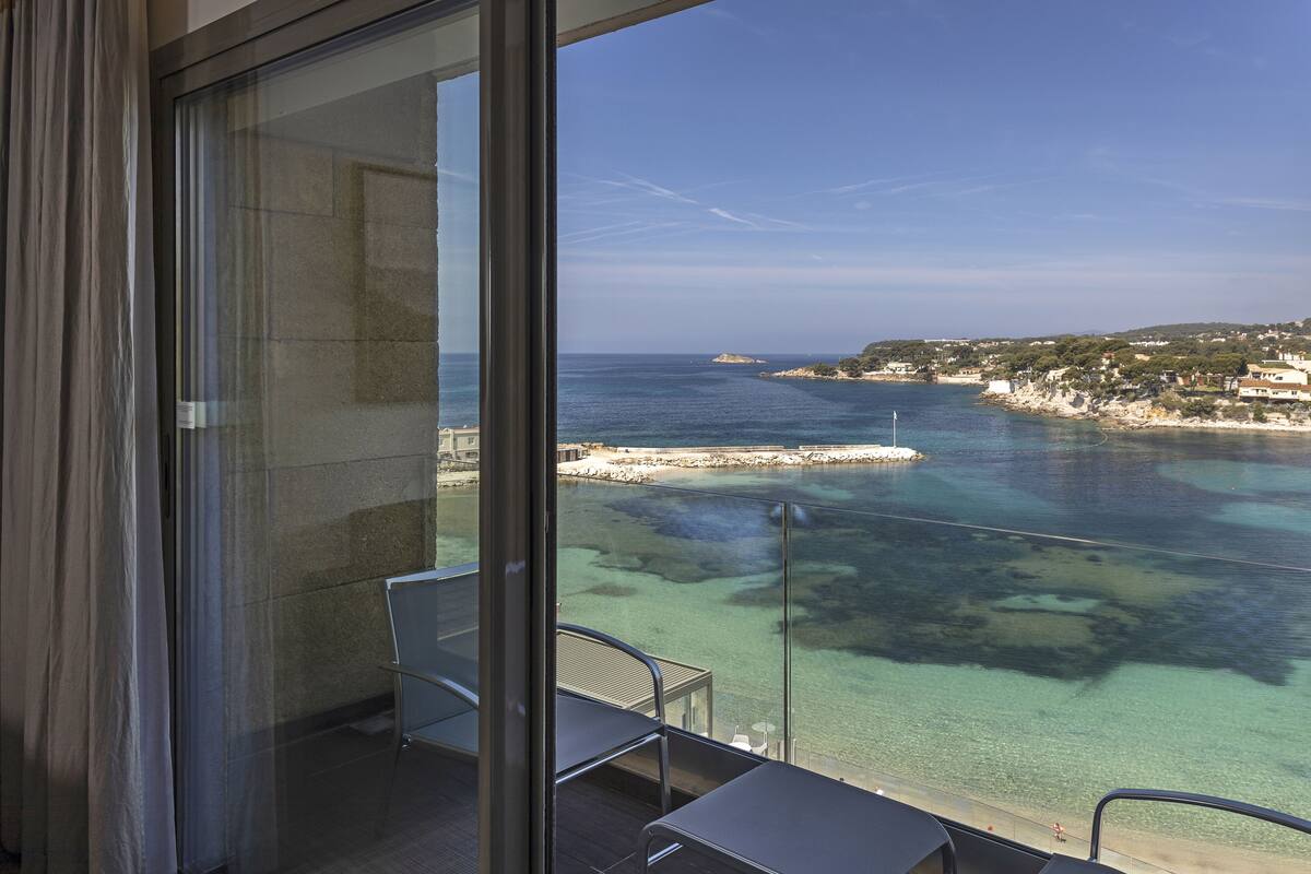 A balcony with a glass door, a table, and two chairs overlooking a coastal area with clear waters and a distant shoreline.