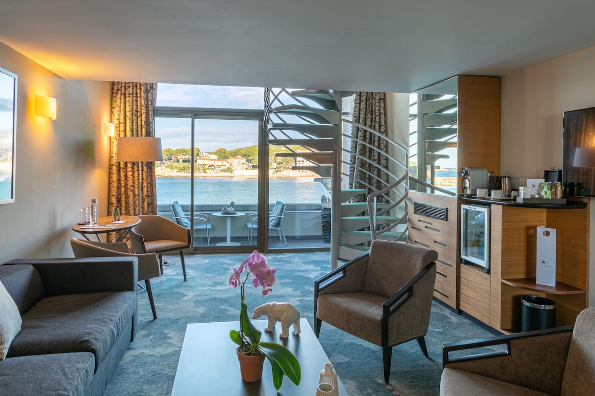 A modern hotel room with a balcony, seating area, and a view of the sea.