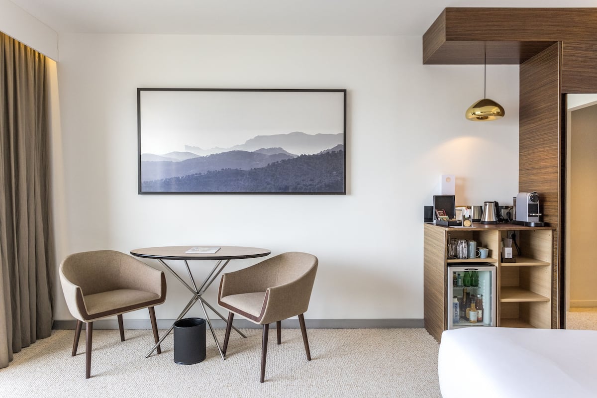 A hotel room with a bed, a desk with a computer, a coffee machine, a small refrigerator, two chairs, a picture on the wall, and a curtain.
