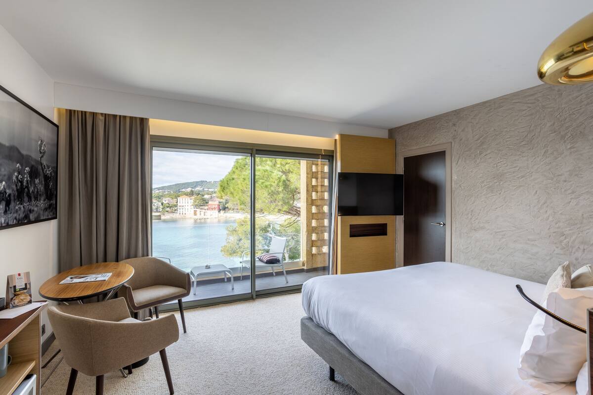 A hotel room with a large bed, a desk with a chair, a TV, and a balcony with a view of the sea.