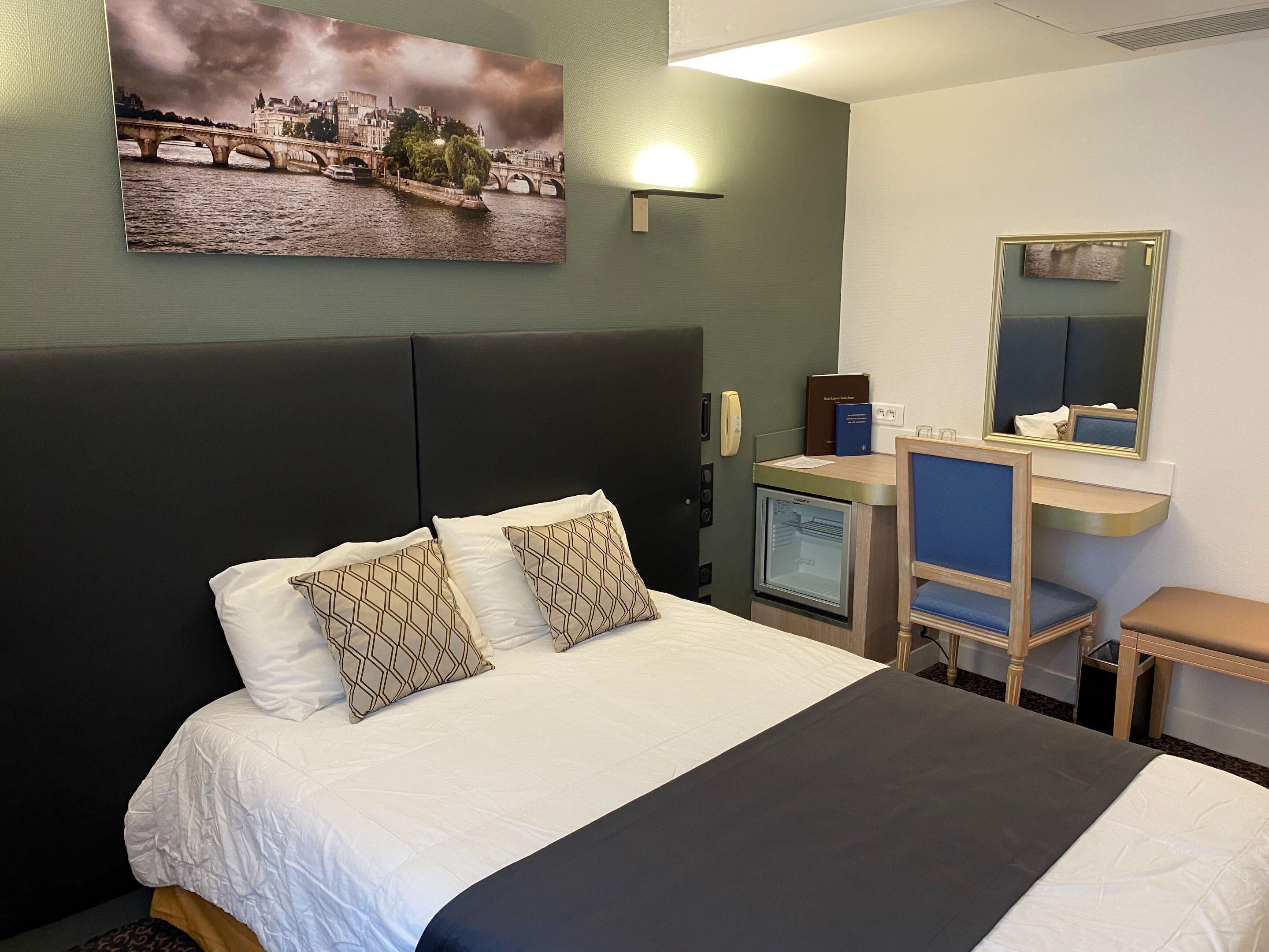 Hotellihuone, jossa on sänky, peilillä varustettu työpöytä, jääkaappi ja maalaus, jossa on kuvattuna silta.