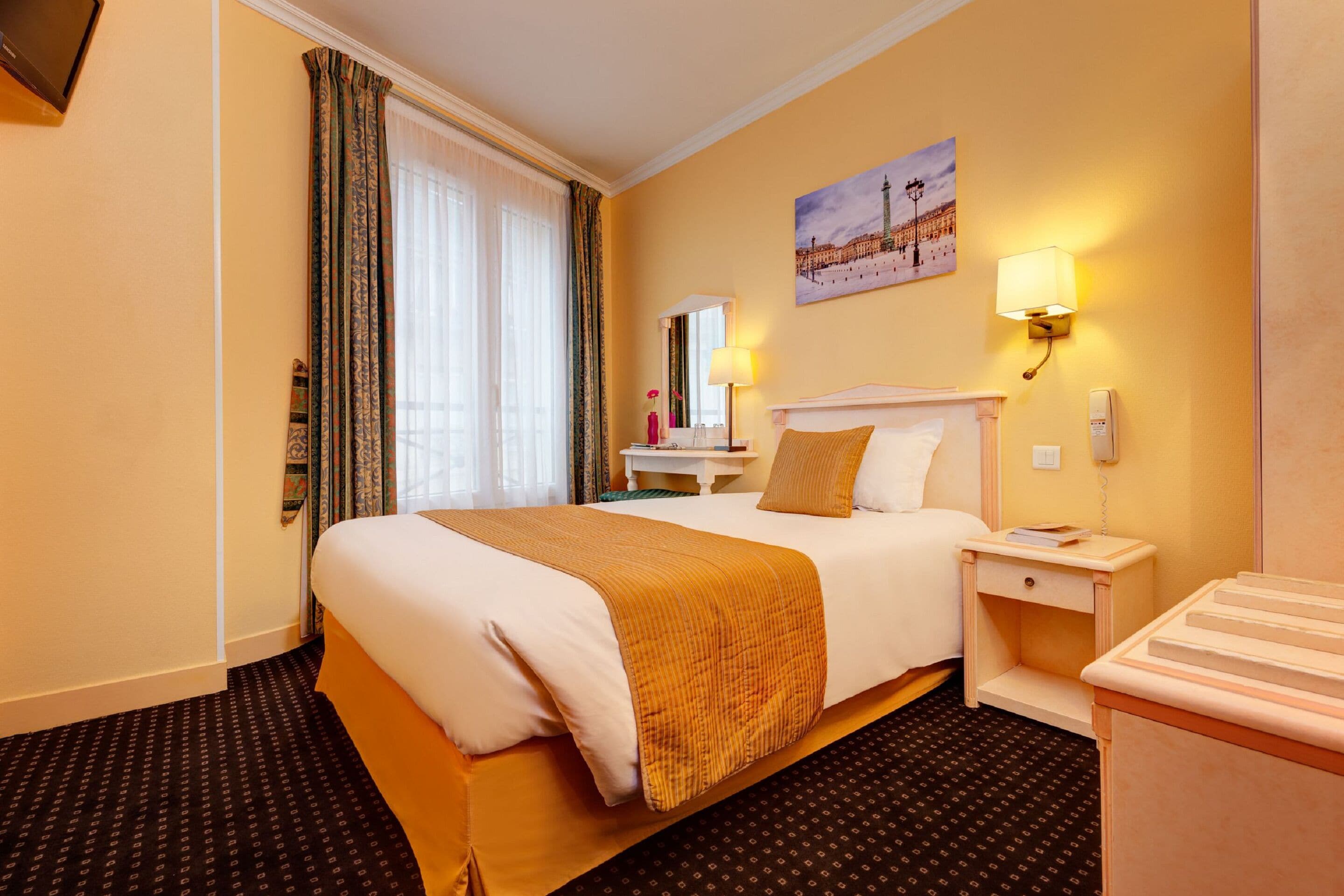 A hotel room with a large bed, a bedside table, a lamp, a painting on the wall, and a carpeted floor.