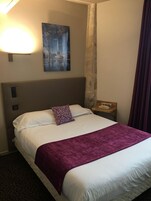 Una camera d'albergo con un letto grande, una testiera, un comodino, una lampada, un quadro appeso al muro e una tenda.