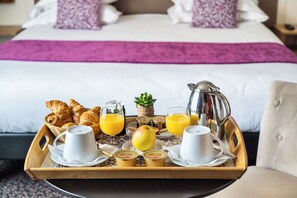 A breakfast tray with croissants, juice, a coffee pot, and a fruit on a bed with a purple bedspread. - Hôtel Louvre Sainte Anne (Paris)