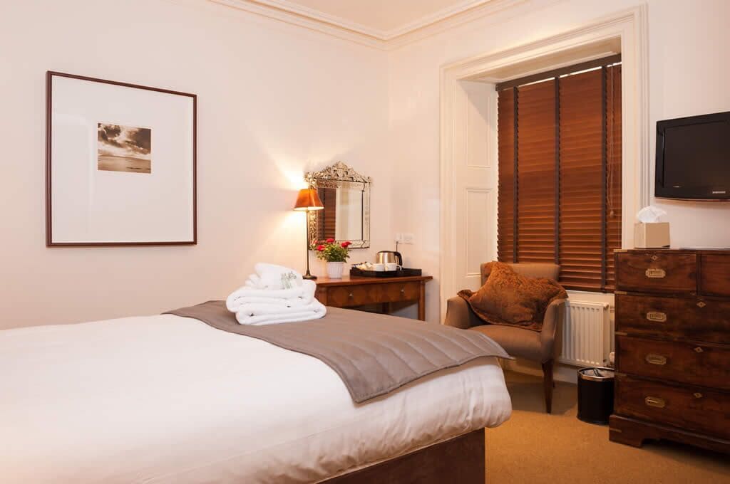Quarto de hotel com cama, penteadeira, cadeira, escrivaninha, espelho, quadro emoldurado e televisão.