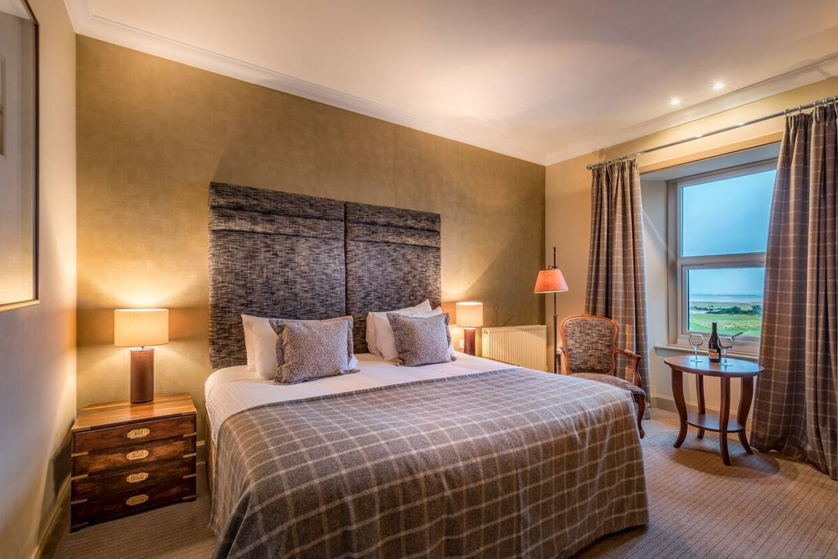 A hotel room with a large bed, two bedside lamps, a chair, a small table, and a view of the sea through the window.