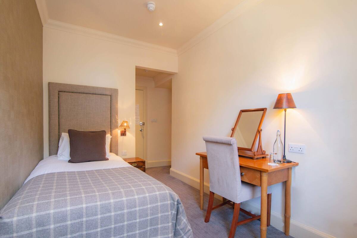 A hotel room with a bed, a desk with a chair, a lamp, and a mirror.