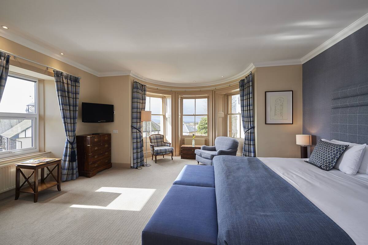 A hotel room with a large bed, two armchairs, a small table, a dresser, and a TV.