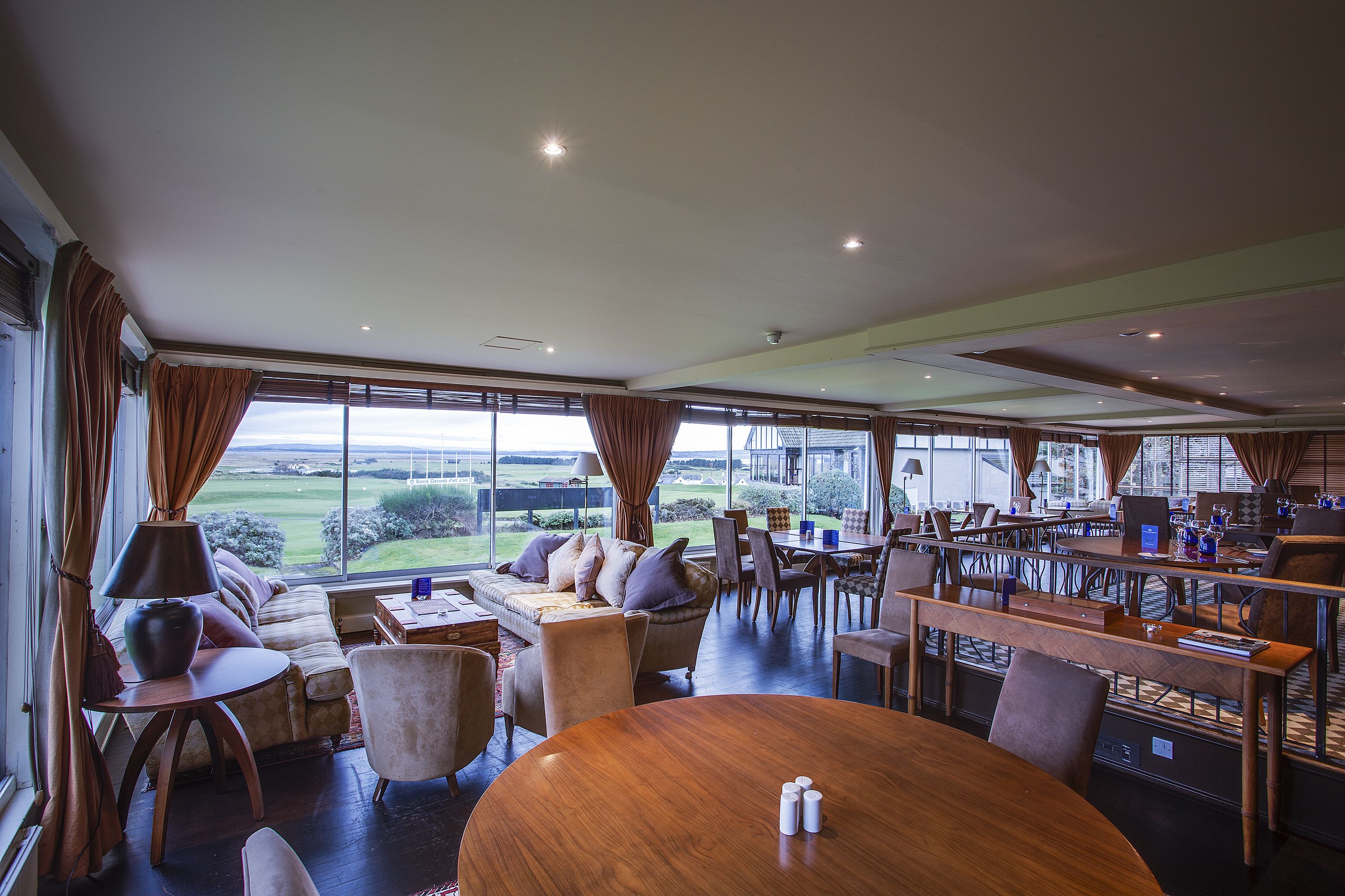 A spacious room with a large window, a round wooden table, and a sofa with cushions.