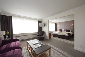 A hotel room with a bed, a chair, a table with a magazine, a TV on the wall, and a window with curtains. - Royal Golf Hotel (Dornoch)