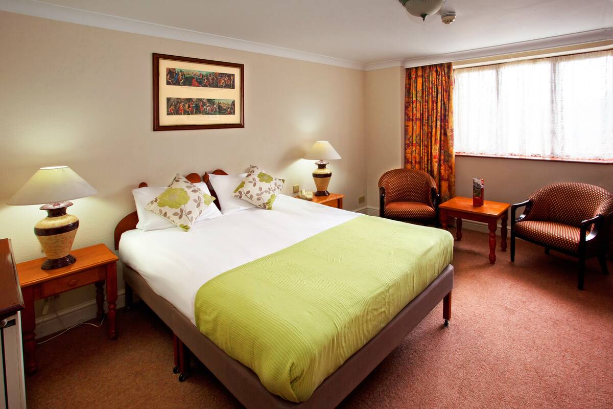A hotel room with a large bed, two bedside tables with lamps, a chair, a window with curtains, and a framed picture on the wall.