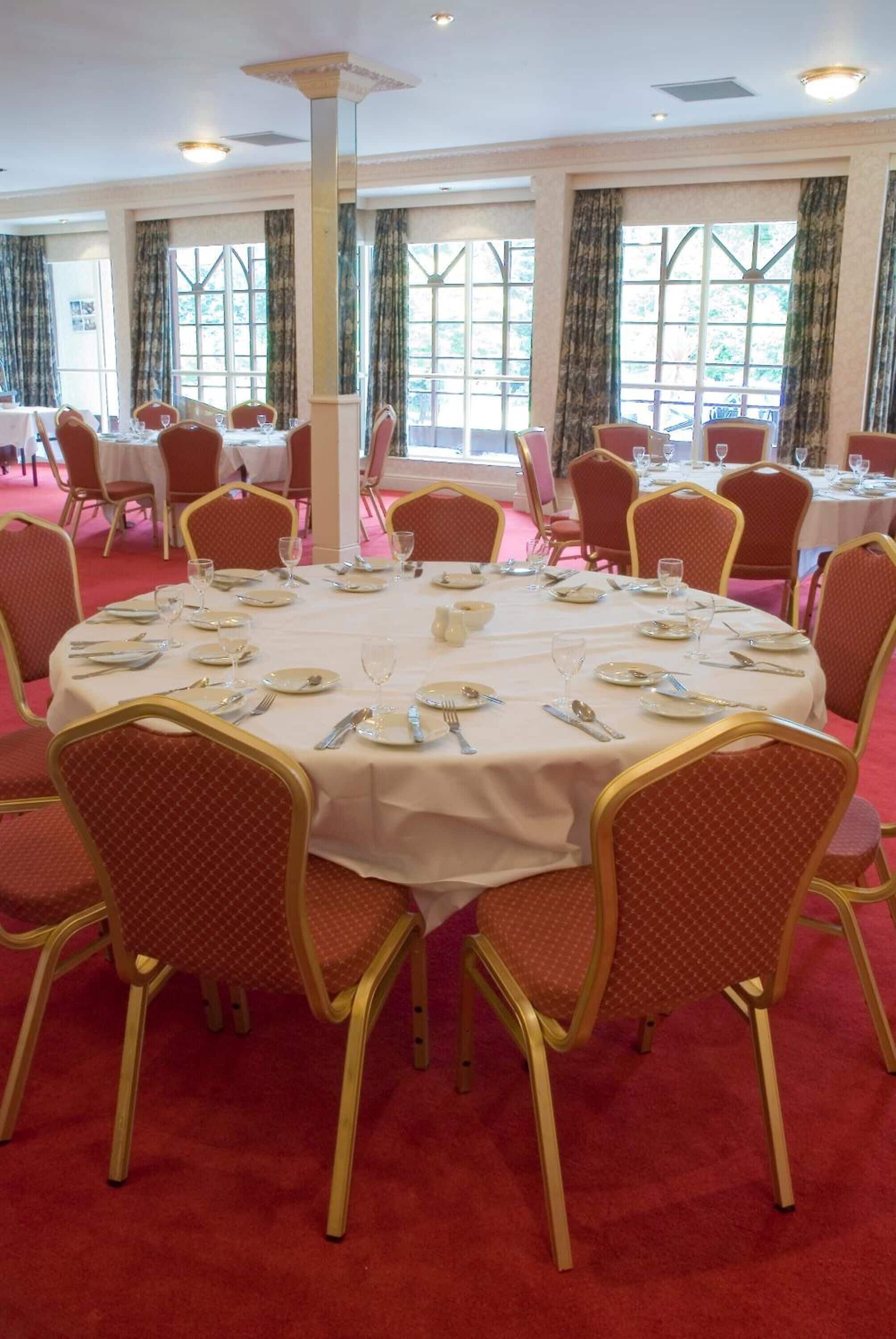 A banquet hall with round tables set for dining, surrounded by red upholstered chairs with gold accents.