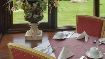 A dining table set for four with a vase of flowers, red upholstered chairs, and a view of a garden and gazebo through large windows.
