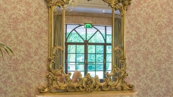 A grand, ornate gold-framed mirror on a marble pedestal, set against a patterned wallpaper backdrop.