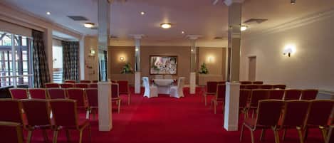 A formal room with red carpet, rows of red chairs, a white table with chairs, and decorative wall sconces.