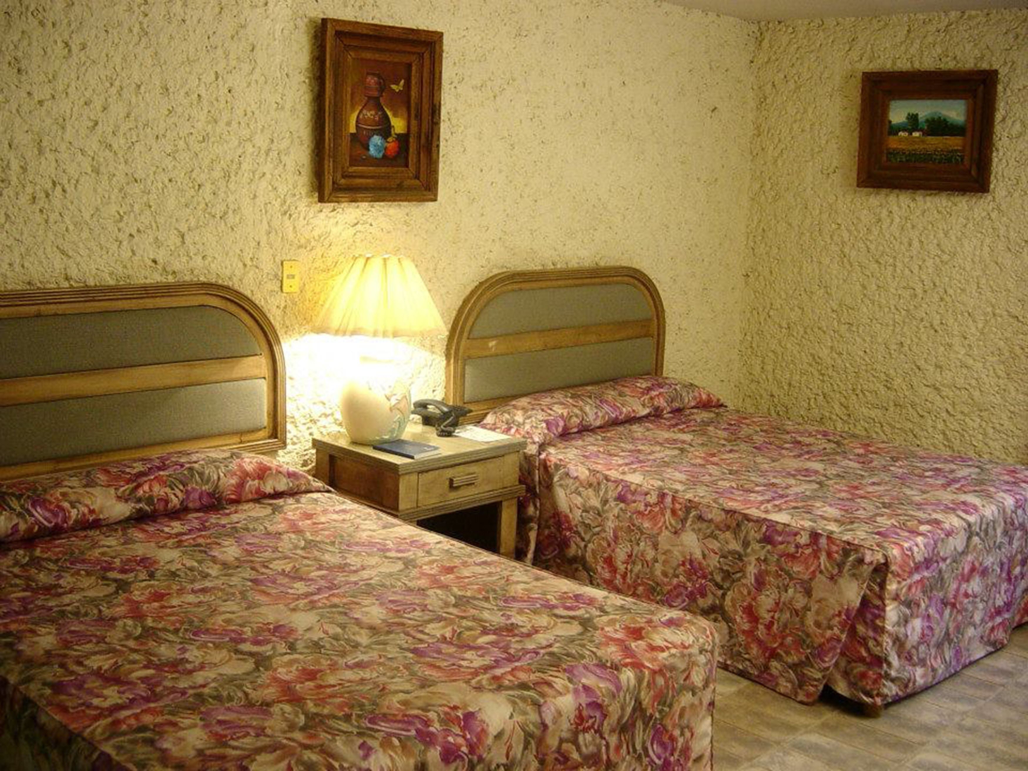 A room with two beds, each with floral bedding, a bedside lamp, and a small table with a lamp.