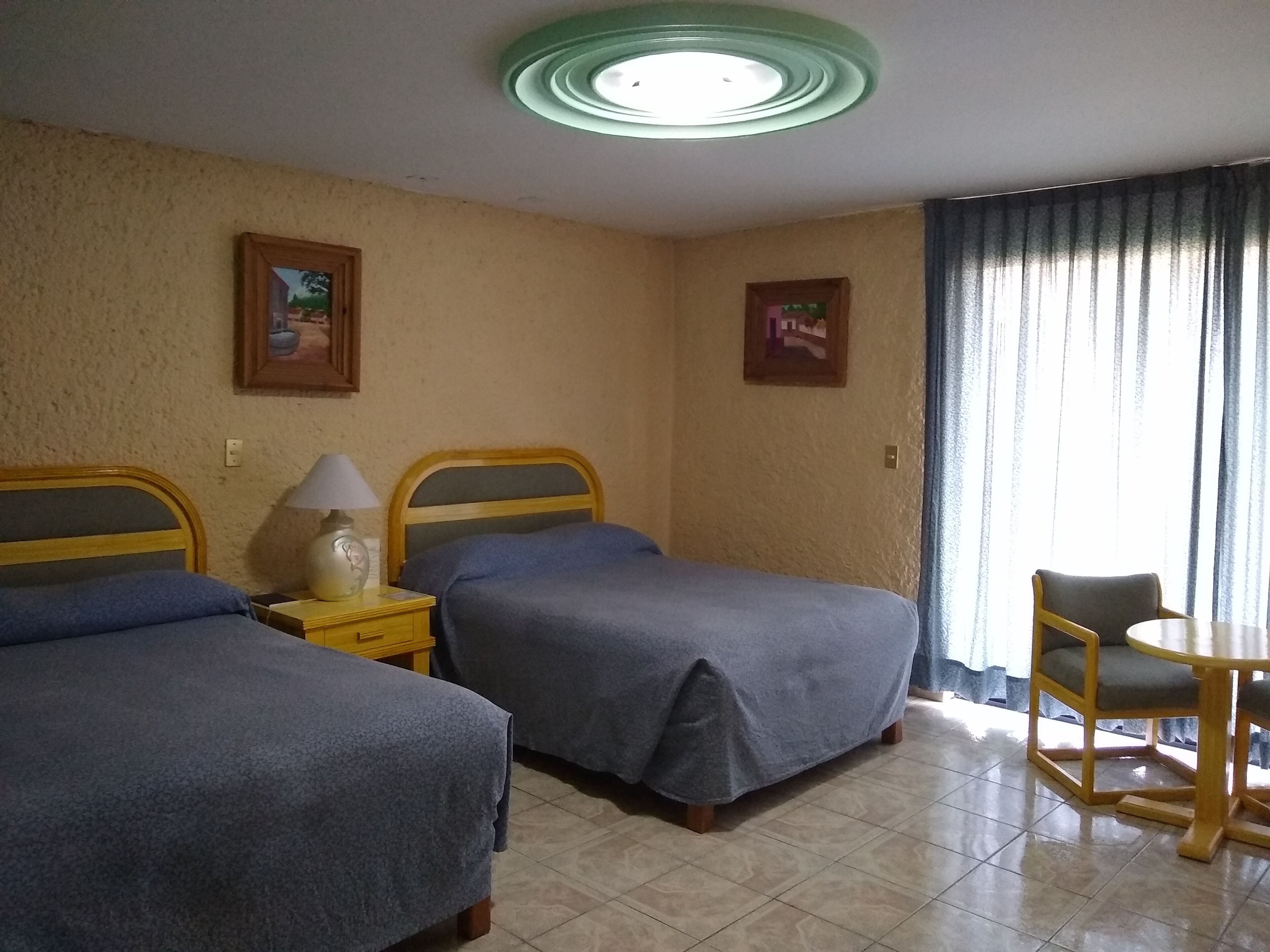 Une chambre d’hôtel avec deux lits simples, une petite table avec une chaise, une lampe et un luminaire au plafond.