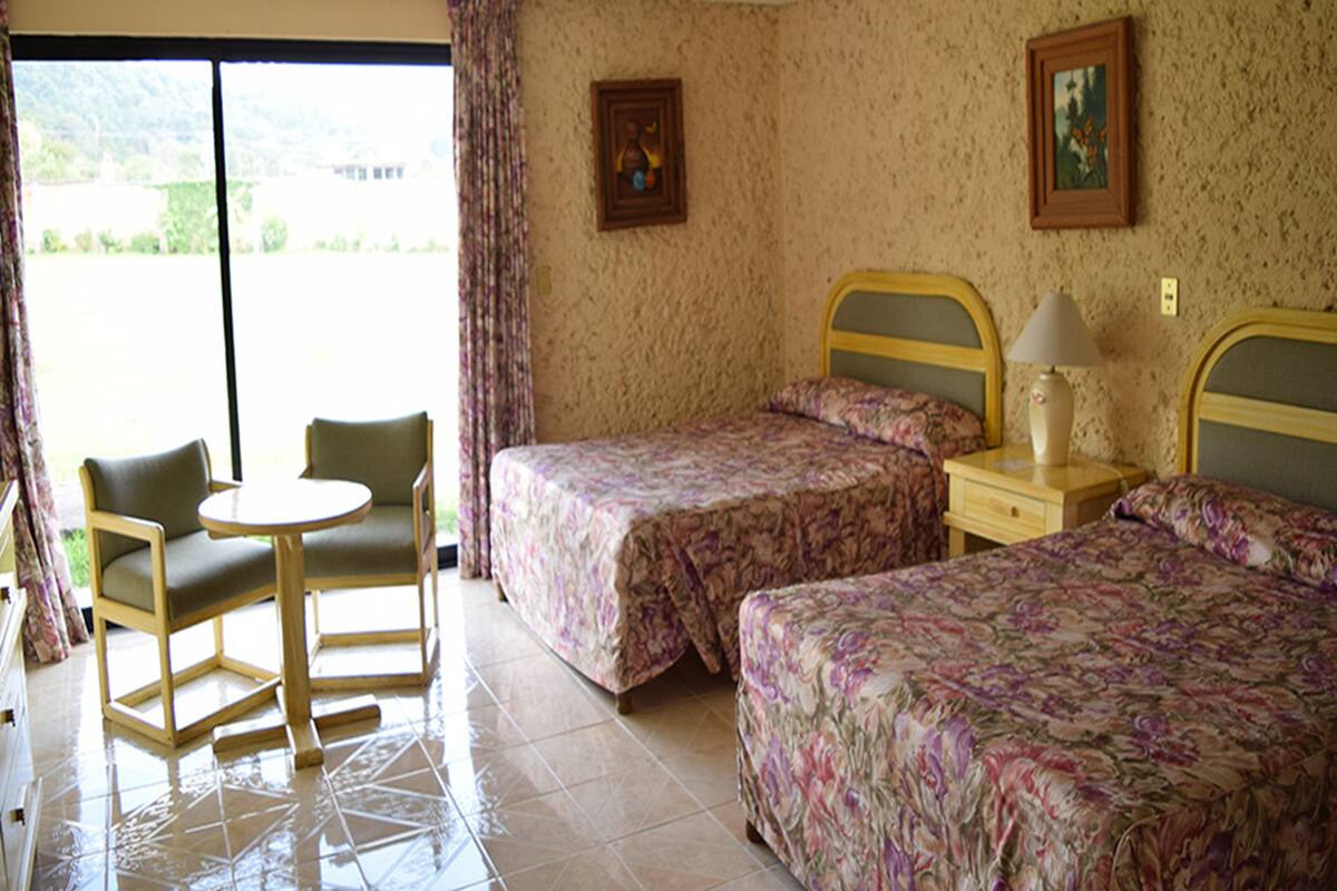 Ein Hotelzimmer mit zwei Betten, einem kleinen Tisch und Stühlen. Es gibt ein Fenster mit Vorhängen, ein Bild an der Wand und eine Lampe auf dem Nachttisch.