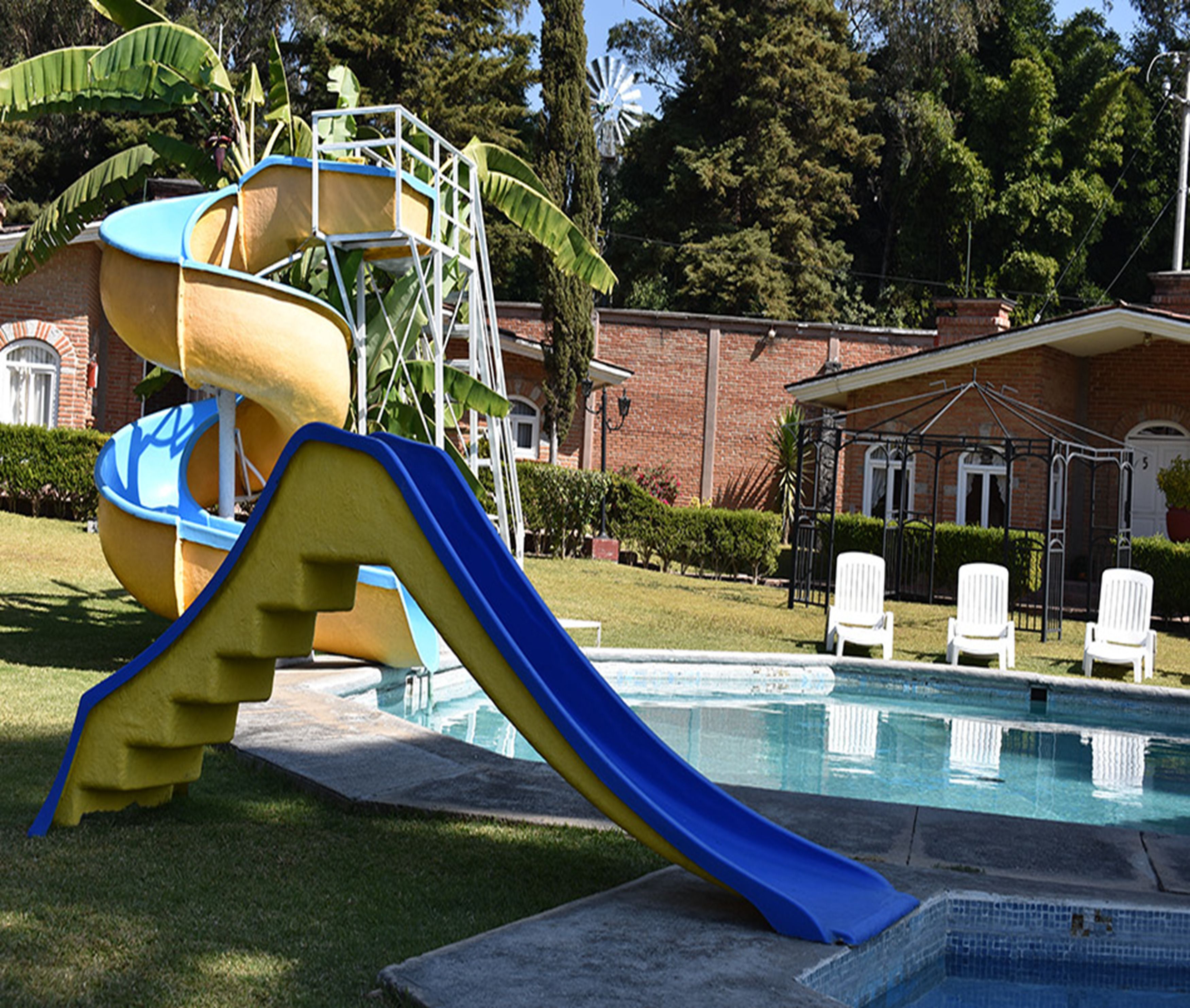 Una piscina con uno scivolo, un edificio in mattoni e sedie bianche sullo sfondo.