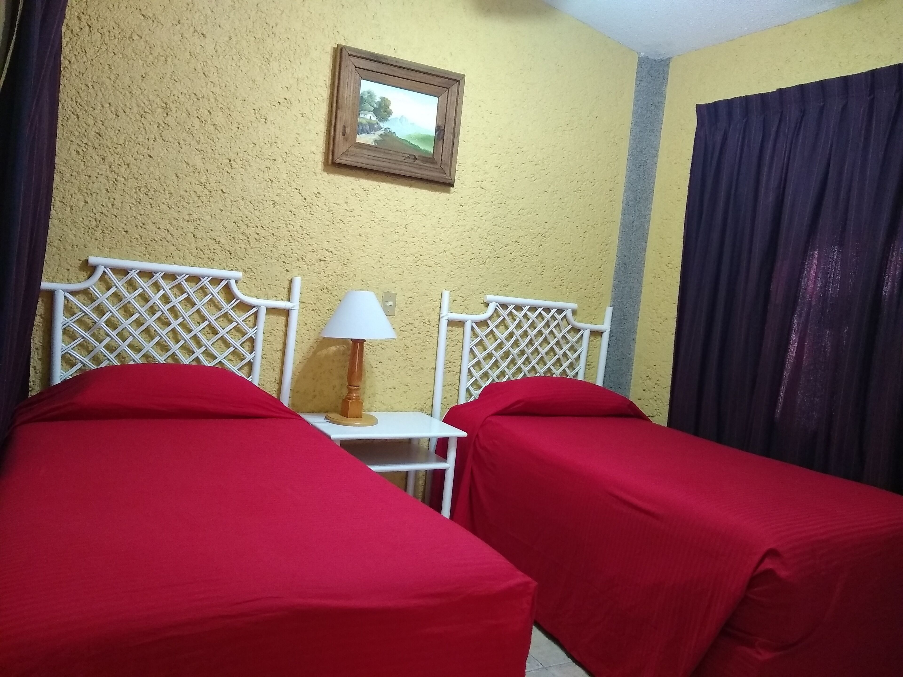 Une chambre avec deux lits, chacun recouvert d’un drap rouge, une table de chevet blanche avec une lampe, et un tableau encadré accroché au mur.