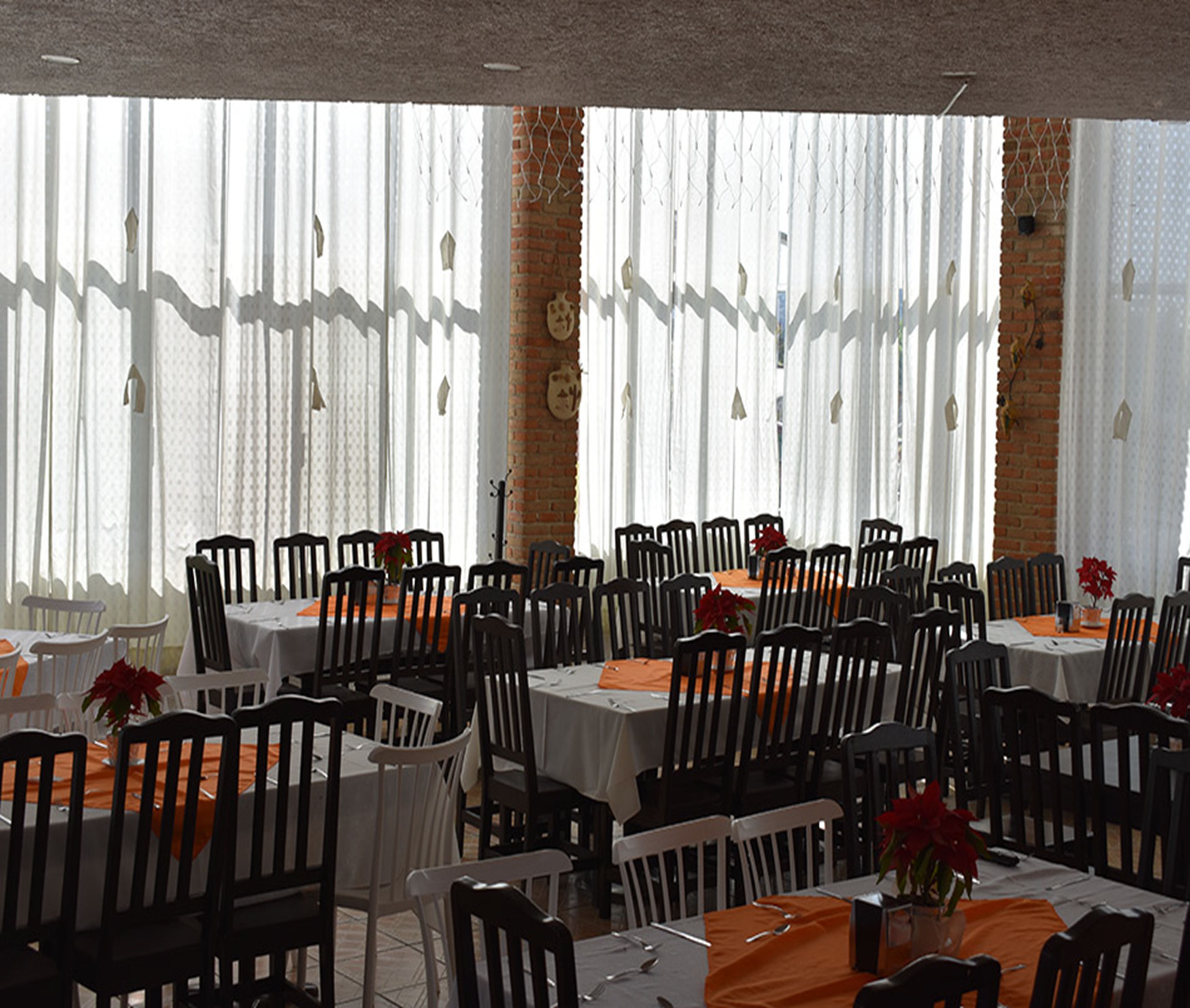 A restaurant with multiple tables and chairs, large windows with sheer curtains, and a brick wall.