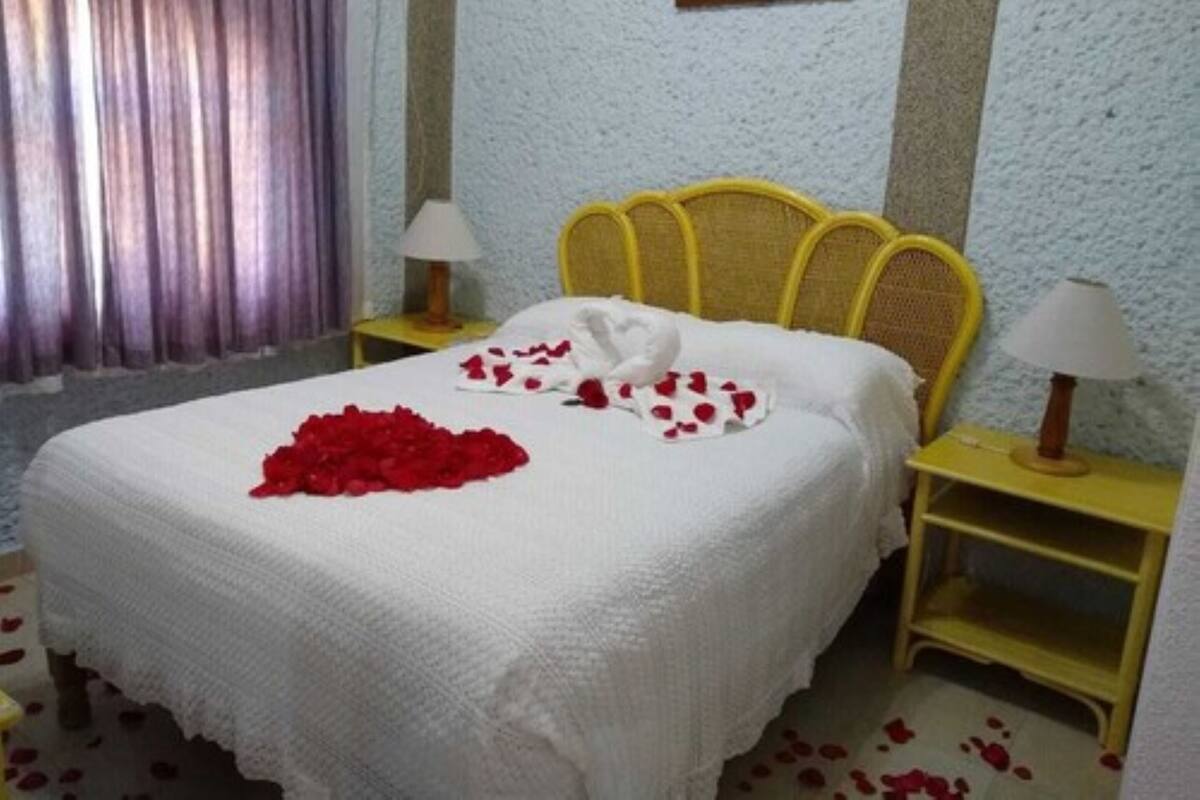 Ein ordentlich bezogenes Bett mit weißer Bettdecke, zwei Kissen und einem Strauß roter Rosen. Zwei Nachttische mit Lampen, ein Fenster mit Vorhängen und ein gerahmtes Bild an der Wand sind ebenfalls zu sehen.