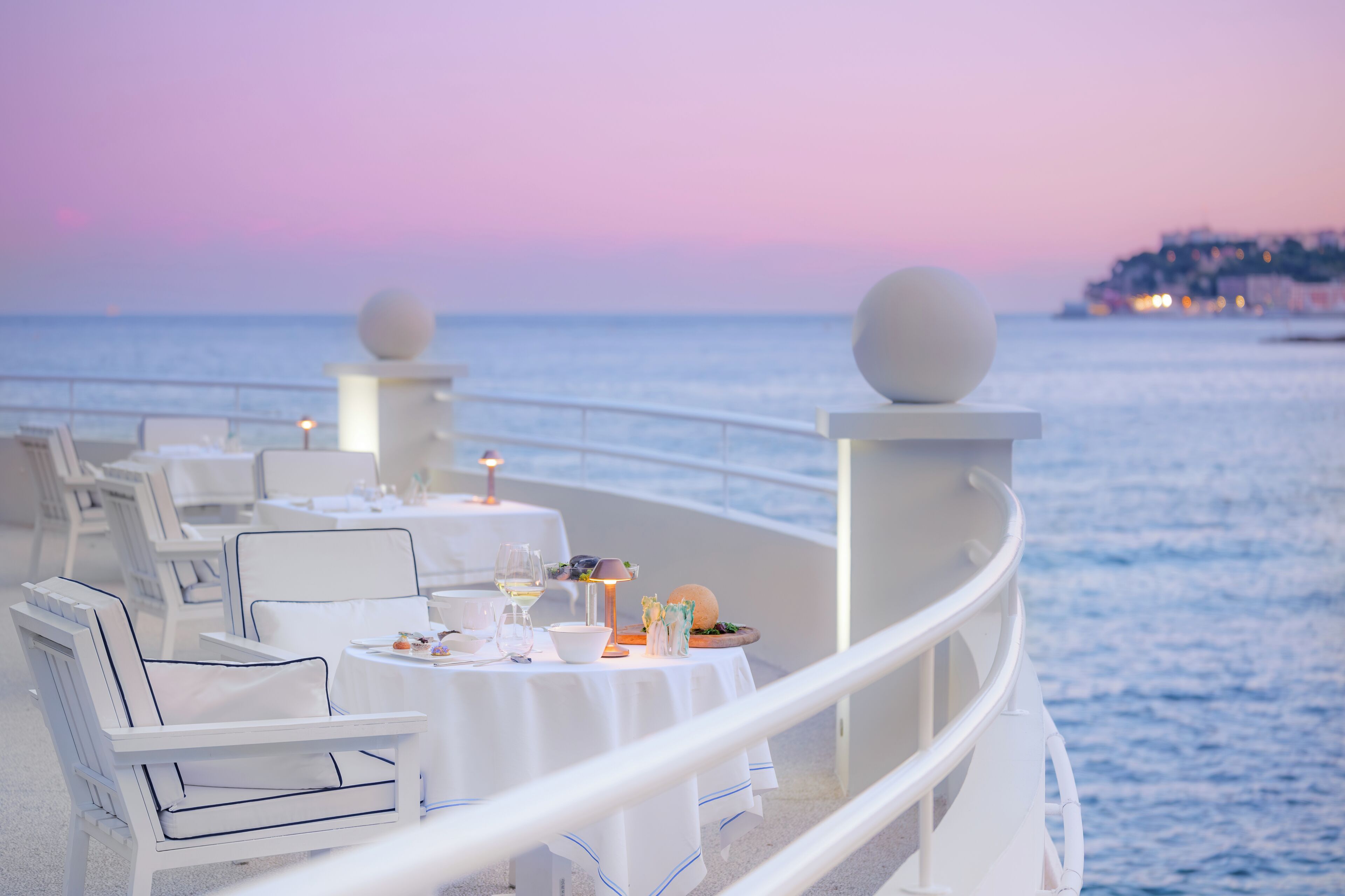 A dining area with white chairs and tables, a view of the sea, and a sunset in the background.