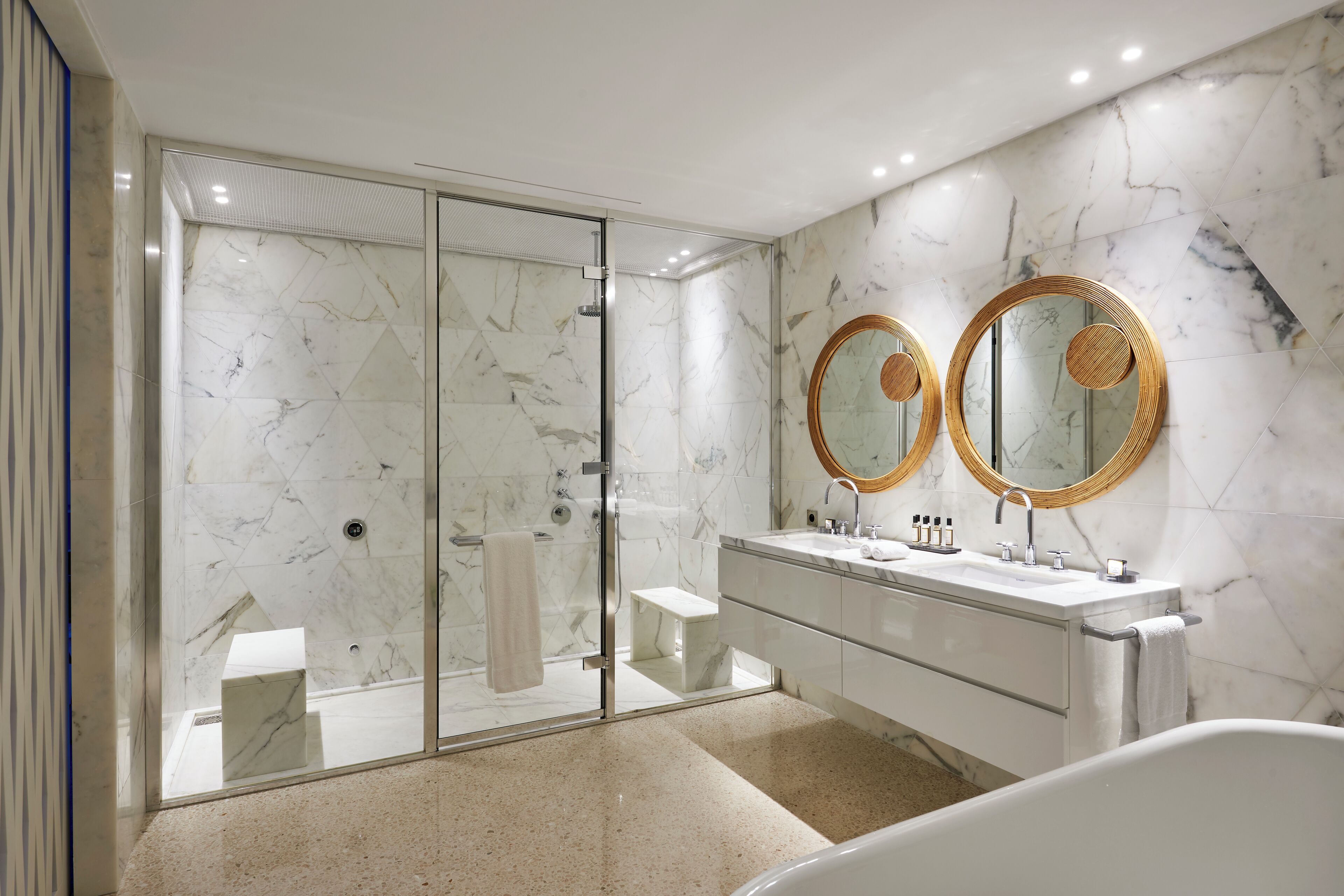 Une salle de bain moderne dotée d’une douche à parois de verre, d’un meuble-lavabo double avec des miroirs à bordure dorée et d’une baignoire sur pieds.