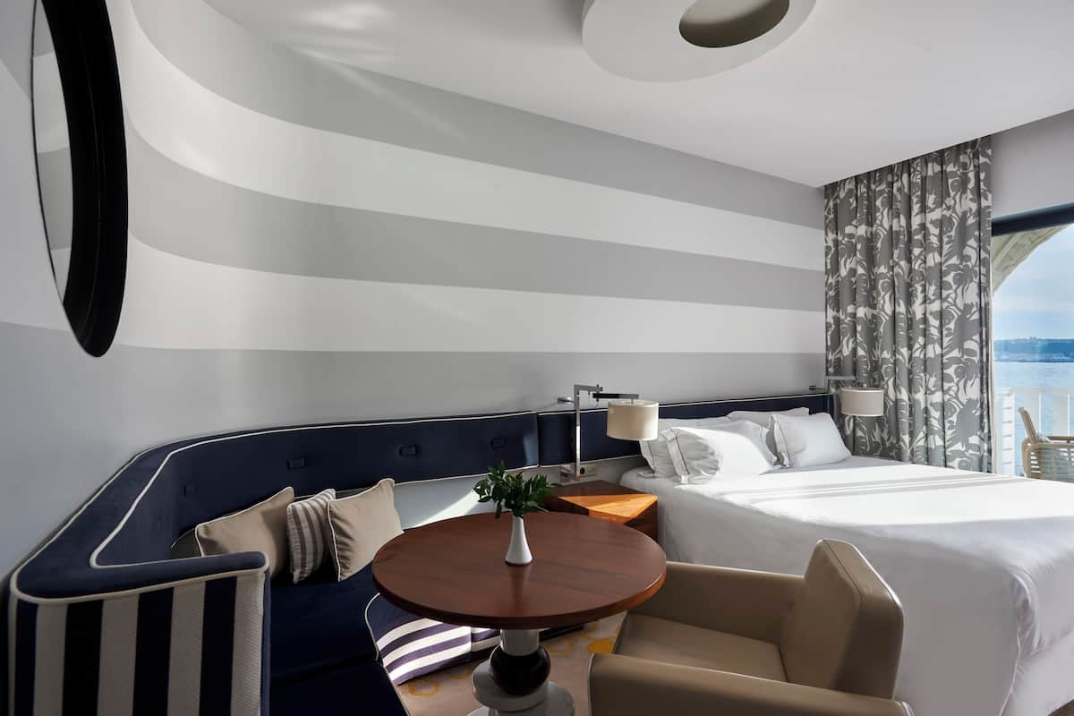 Une chambre d’hôtel moderne avec un lit aux lignes courbes, une table ronde agrémentée d’une plante, un canapé et une vue sur la mer.