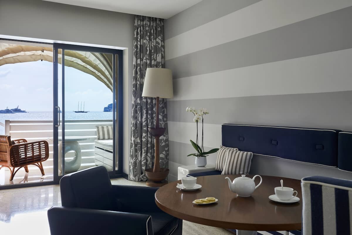 Une chambre d’hôtel moderne avec un balcon, une table ronde agrémentée d’un service à thé, une orchidée en pot et un rideau à rayures.