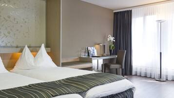 Ein Hotelzimmer mit einem großen Bett, einem Schreibtisch mit Stuhl, einer Stehlampe und Blick ins Freie durch ein mit Vorhängen versehenes Fenster.